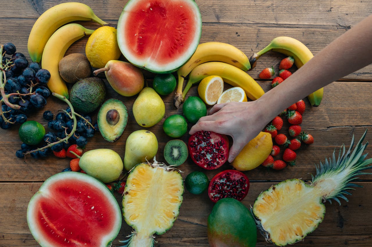 Colorful variety of fresh fruits arranged on a wooden surface with a hand selecting a pomegranate.